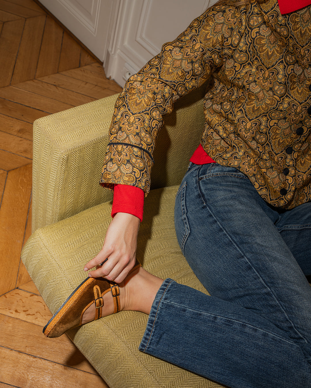 Woman on a green sofa adjusting the buckle of the Nocciola Double Mary Jane, worn with jeans
