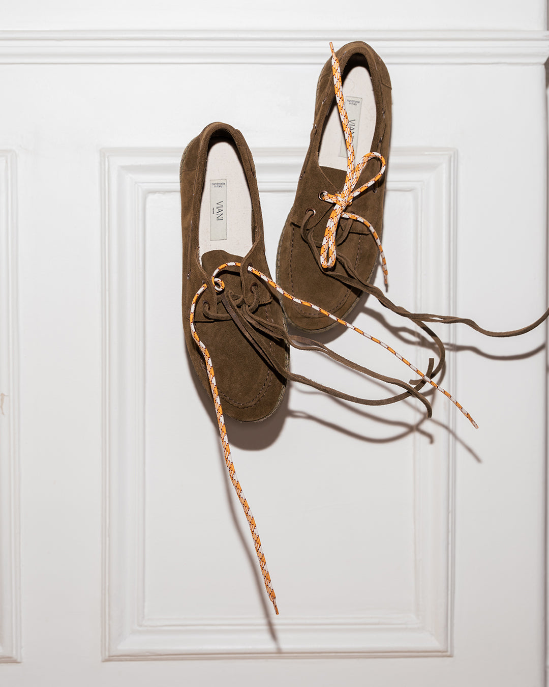Pair of Olive Scarpa da Barca hanging by their suede laces against a white door