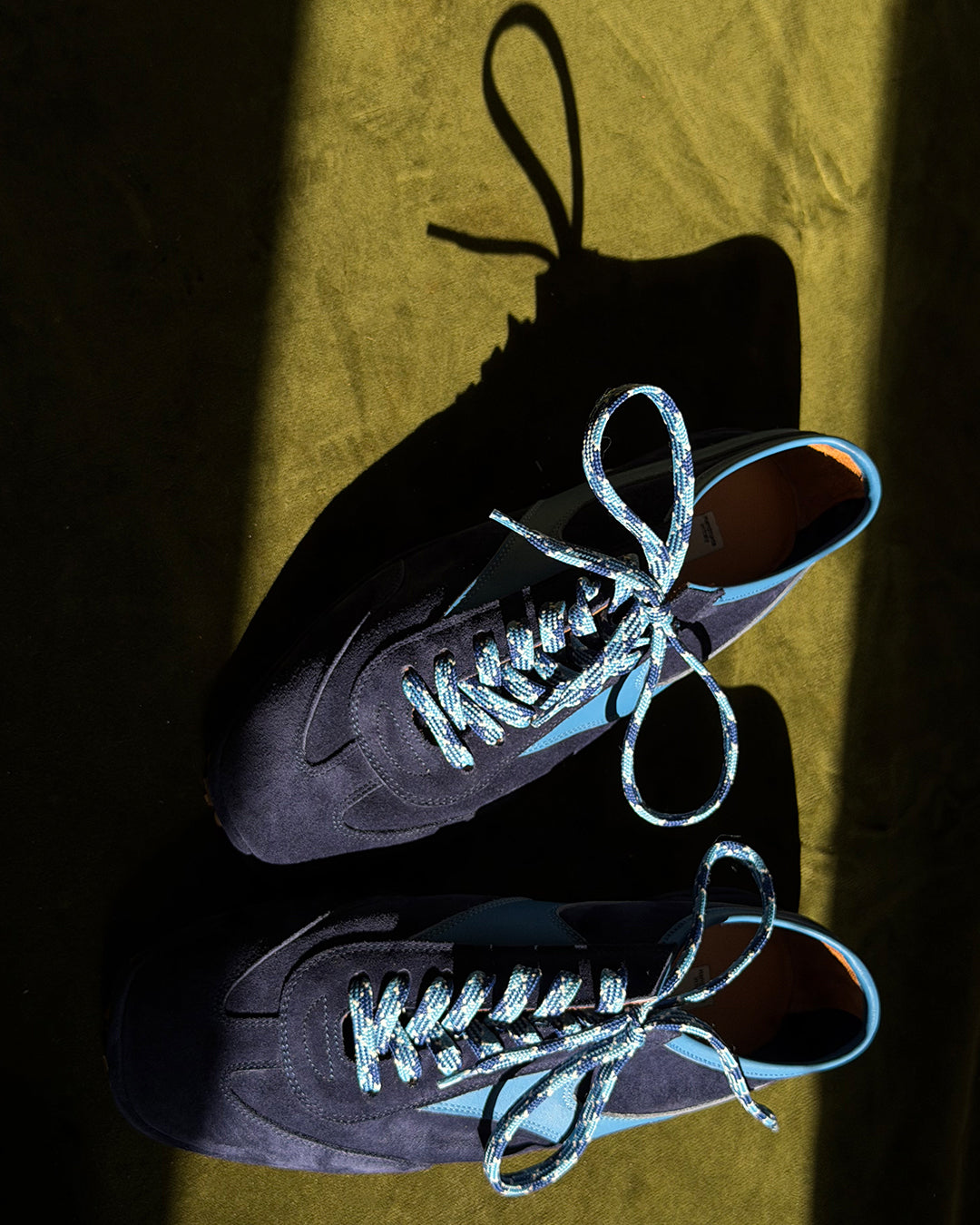 Pair of Trieste Corsa Sneakers in navy suede with blue braided laces, top view in sunlight